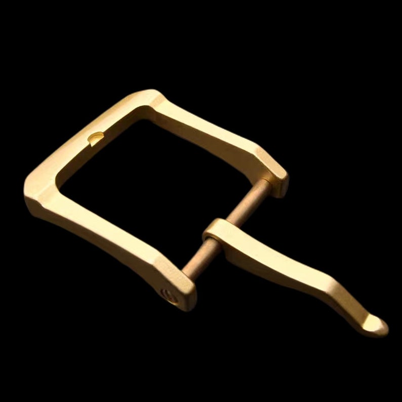 May include: A gold-toned metal buckle with a rectangular frame and a curved, hinged clasp. The buckle is designed for a strap or belt.