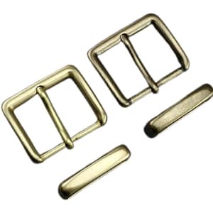 Könnte beinhalten: Zwei messingfarbene Gürtelschnallen und zwei passende Gürtelschlaufen. Die Schnallen sind rechteckig mit einem einzelnen Steg, und die Schlaufen sind abgerundete Rechtecke. Die Hardware ist neu und einsatzbereit.