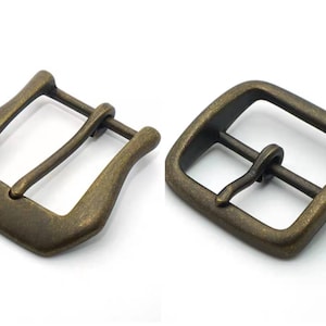 Pode incluir: Duas fivelas de cinto de metal de cor dourada antiga. Uma fivela é de forma retangular tradicional, a outra é mais arredondada, de forma oval.