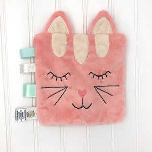 May include: Pink plush cat security blanket with embroidered eyes, nose, and whiskers. The blanket has three satin ribbons attached to the side.