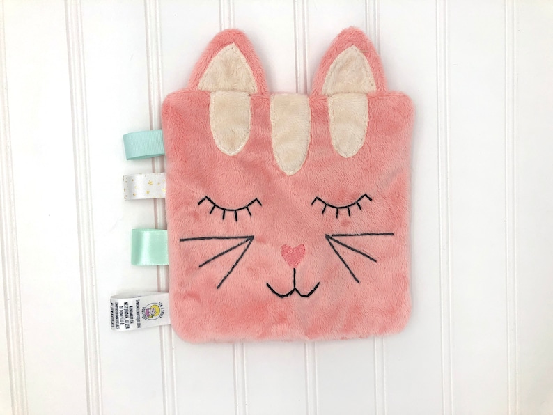 May include: Pink plush cat security blanket with embroidered eyes, nose, and whiskers. The blanket has three satin ribbons attached to the side.
