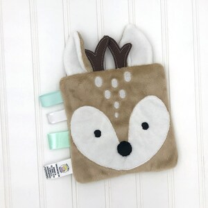 May include: A brown and white plush deer-shaped baby toy with a white face and black eyes. The toy has a soft, fuzzy texture and is attached to a set of three satin ribbons in shades of green and white.