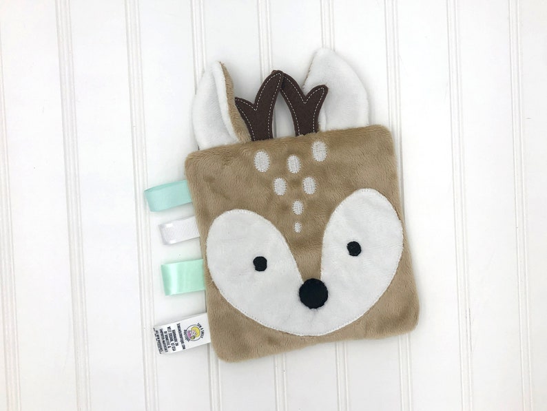 May include: A brown and white plush deer-shaped baby toy with a white face and black eyes. The toy has a soft, fuzzy texture and is attached to a set of three satin ribbons in shades of green and white.