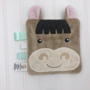 May include: A brown plush horse with pink ears and a white face. The horse has a stitched smile and black eyes. The plush toy has three satin ribbons attached to one side.