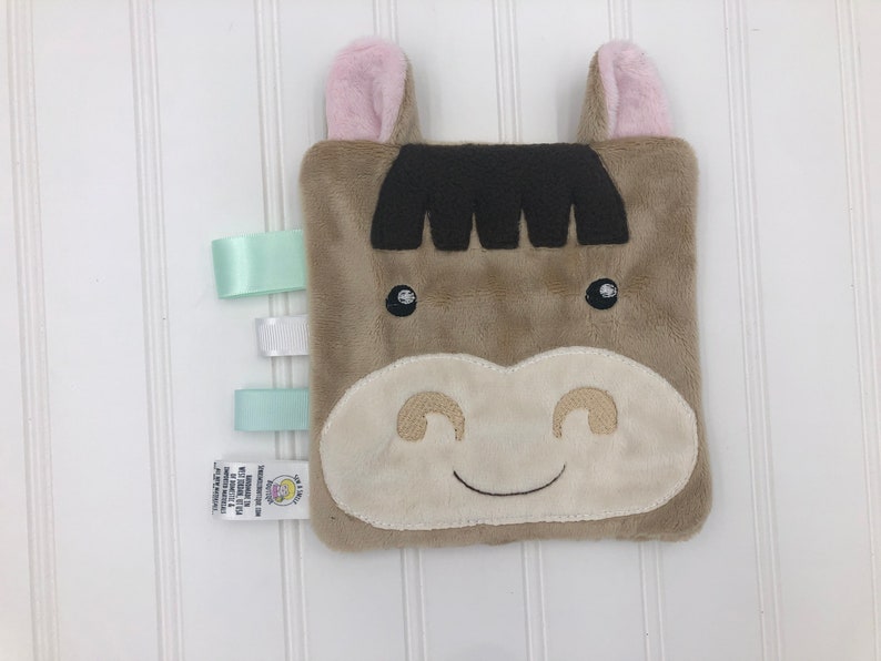 May include: A brown plush horse with pink ears and a white face. The horse has a stitched smile and black eyes. The plush toy has three satin ribbons attached to one side.