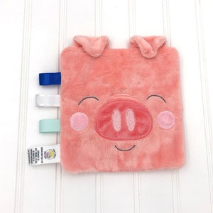 May include: A pink plush pig security blanket with a smiling face and embroidered details. It has three fabric tags attached to the side: blue, white, and light blue.
