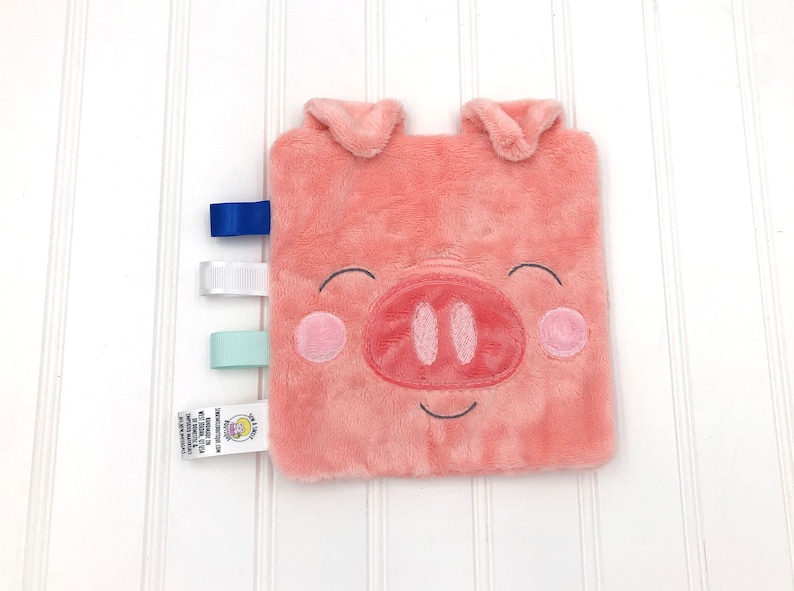 May include: A pink plush pig security blanket with a smiling face and embroidered details. It has three fabric tags attached to the side: blue, white, and light blue.