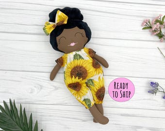 May include: A handmade doll with brown skin, black hair, and a sunflower print dress. The doll is smiling and has a pink bow in her hair. The doll is lying on a white wooden surface. "Ready to Ship" is written on a pink circle.