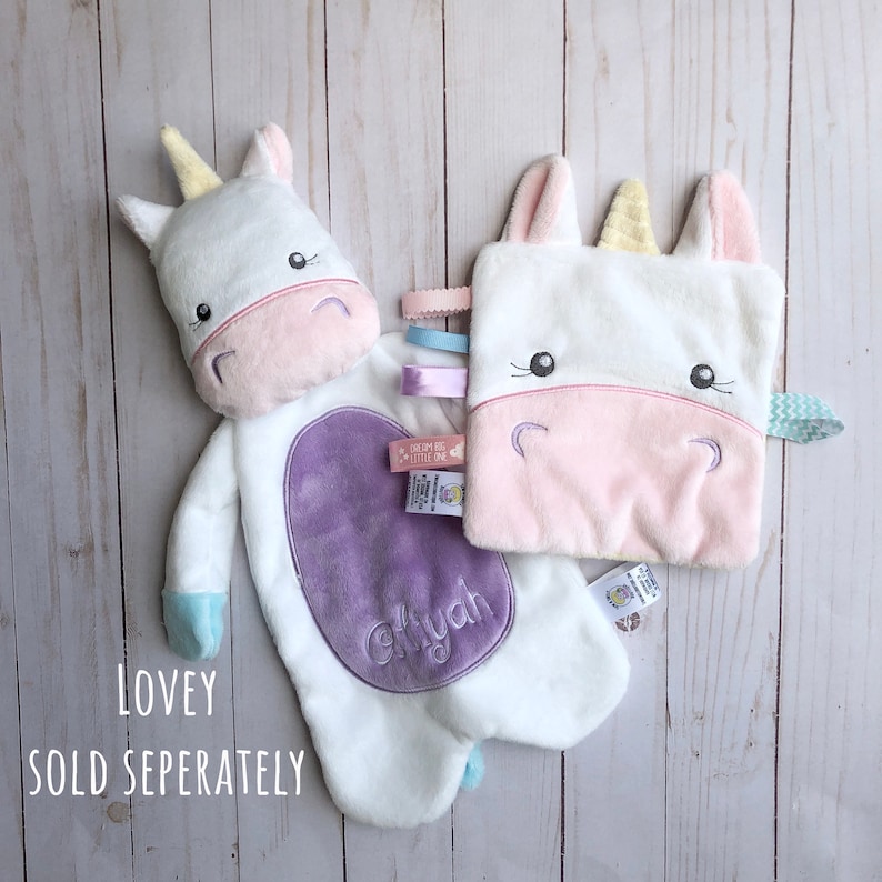 May include: Two white unicorn loveys with embroidered faces and pastel colored details. One lovey is a plush toy with a purple oval patch that says "Aaliyah". The other lovey is a smaller square with a pink face and a rainbow ribbon.