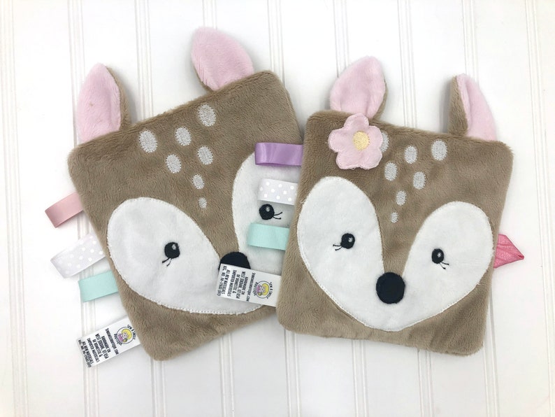May include: Two brown plush fawn baby security blankets with pink ears and embroidered faces. One blanket has a pink flower and the other has a light blue, white, and purple ribbon.