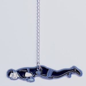 May include: A black and grey enamel pin showing a person in a push-up position. The pin is attached to a silver chain and a magnetic backing. The person wears glasses and a black outfit with grey accents.