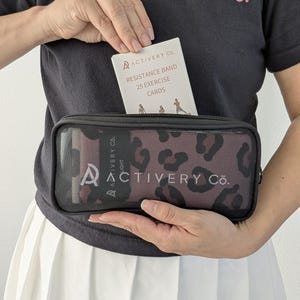 May include: A black zippered case with a leopard print pattern, containing resistance bands and exercise cards. The exercise cards are labelled "Resistance Band 25 Exercise Cards". The case is held in front of a black t-shirt with the text "ACTIVERY Co."
