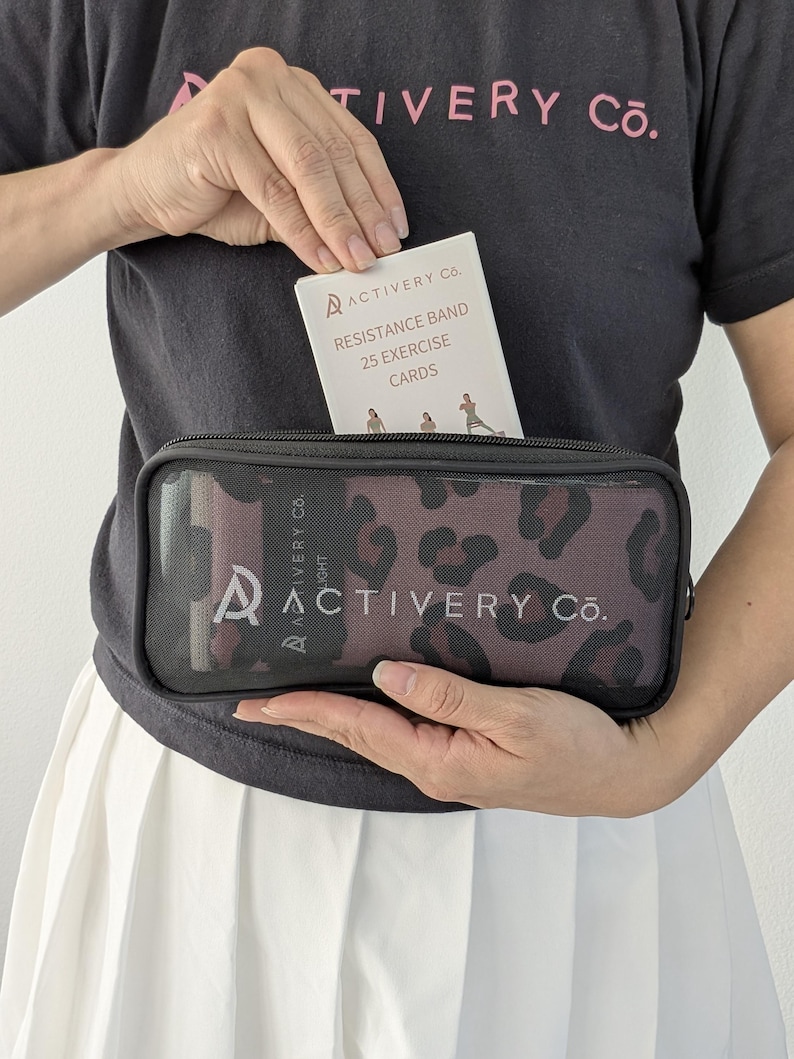 May include: A black zippered case with a leopard print pattern, containing resistance bands and exercise cards. The exercise cards are labelled "Resistance Band 25 Exercise Cards". The case is held in front of a black t-shirt with the text "ACTIVERY Co."