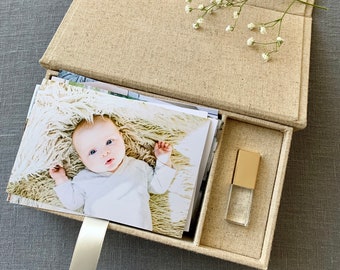 May include: A beige linen box with a white ribbon, containing a stack of photos of a baby and a small gold and clear USB drive. The box is open and there are white flowers on top.