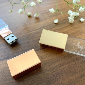 May include: Three gold and clear USB flash drives. Two of the drives are rectangular and have a gold metal finish. The third drive is rectangular and made of clear glass with a gold metal cap. The glass drive has the initials "hg" engraved on it.