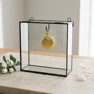 Op de afbeelding: Een rechthoekige glazen displaybox met een dun zwart metalen frame. Binnenin hangt een goudkleurig zakhorloge aan een klein haakje dat aan het bovenste frame is bevestigd. De box heeft twee kleine ronde lussen aan de bovenrand.