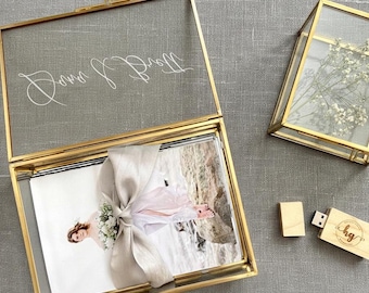 May include: A rectangular glass box with a gold metal frame featuring the white script text Dana & Brett, containing a photograph tied with a beige ribbon. A smaller square glass box with a gold frame and a wooden USB drive with the logo hg.