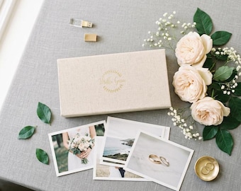 May include: A rectangular beige linen box with the gold embossed text Mellie Grace, accompanied by three printed photographs, a gold USB drive, a small gold dish with two rings, and a cluster of pale pink roses with green leaves.
