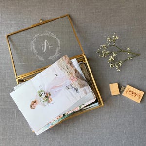 May include: A rectangular glass display box with a gold metal frame and a hinged lid featuring an etched letter A inside a floral wreath. The box contains a stack of photographs. Beside the box is a small wooden USB drive and a sprig of baby's breath.