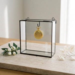 May include: A rectangular glass display case with a thin black metal frame and two top loops. Inside, a gold pocket watch hangs from a central hook. A white etched cross design is visible on the front glass panel of the display box.