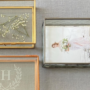 May include: Three glass display boxes with gold and copper frames. One box contains dried white flowers, another a photo of a woman in a pink dress, and the third has a white "H" and laurel wreath design on a gray background.