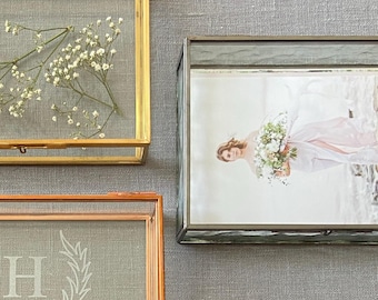 May include: Three rectangular glass display boxes with metal frames. One gold frame contains dried white flowers, one copper frame features a white letter H and laurel wreath design, and one silver frame holds a photograph of a person with flowers.