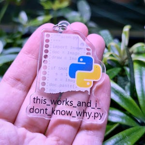 May include: A clear acrylic keychain shaped like a torn piece of paper with Python code printed on it. The code includes the text "this_works_and_i_dont_know_why.py". A yellow and blue Python logo is on the right side of the keychain.