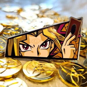 May include: A gold and black enamel pin featuring a close-up of the anime character Yugi Mutou from the Yu-Gi-Oh! franchise. Yugi is shown with his signature spiky blond hair, red eyes, and the Egyptian God Eye symbol on his forehead. He is holding a playing card with the text "Magic" on it.