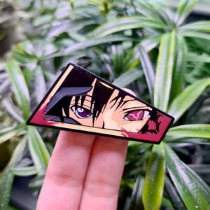 Puede incluir: Un pin de esmalte triangular con fondo negro y rojo. El pin presenta un primer plano de un ojo de un personaje de anime con un iris morado y una pupila roja. El cabello del personaje es negro y su rostro está parcialmente oscurecido por una sombra.