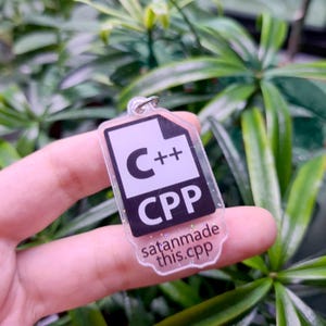May include: A clear plastic keychain with glitter embedded in the plastic. The keychain features a white file icon with the text "C++" and "CPP" in black. The text "satanmade this.cpp" is printed in black at the bottom of the keychain.