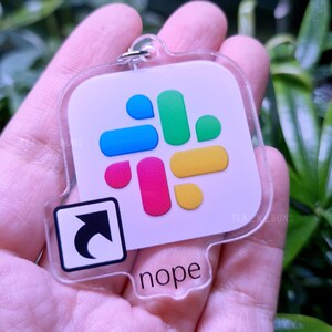 Peut inclure: Un porte-clés en acrylique transparent avec un logo Slack coloré et le mot "nope" imprimé en dessous. Le porte-clés comprend également une flèche noire pointant vers la gauche.
