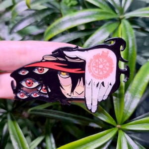 May include: A black and white enamel pin featuring a character with multiple red eyes and a red headband. The character is holding a hand with a pentagram symbol in the center.