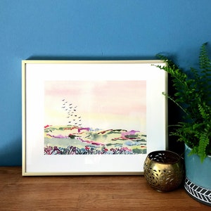 Puede incluir: Una pintura de acuarela enmarcada de un paisaje con una bandada de p&aacute;jaros volando sobre colinas onduladas. La pintura tiene un cielo rosa y amarillo con colinas verdes y moradas. El marco es de madera clara.