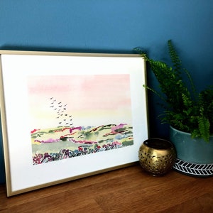 Puede incluir: Una pintura de acuarela enmarcada de un paisaje con un cielo rosa y amarillo, colinas verdes y una bandada de p&aacute;jaros volando por encima.