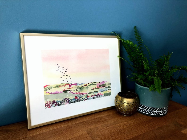 Puede incluir: Una pintura de acuarela enmarcada de un paisaje con un cielo rosa y amarillo, colinas verdes y una bandada de p&aacute;jaros volando por encima.