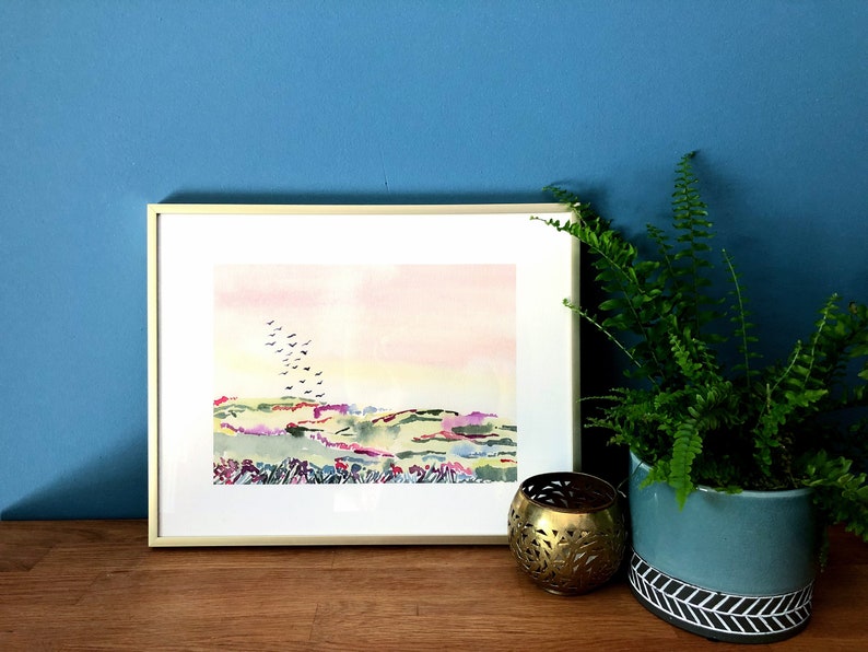 Puede incluir: Una pintura de acuarela enmarcada de un paisaje con una bandada de p&aacute;jaros volando sobre colinas onduladas. La pintura tiene un cielo rosa y amarillo con colinas verdes y moradas. El marco es de madera clara.