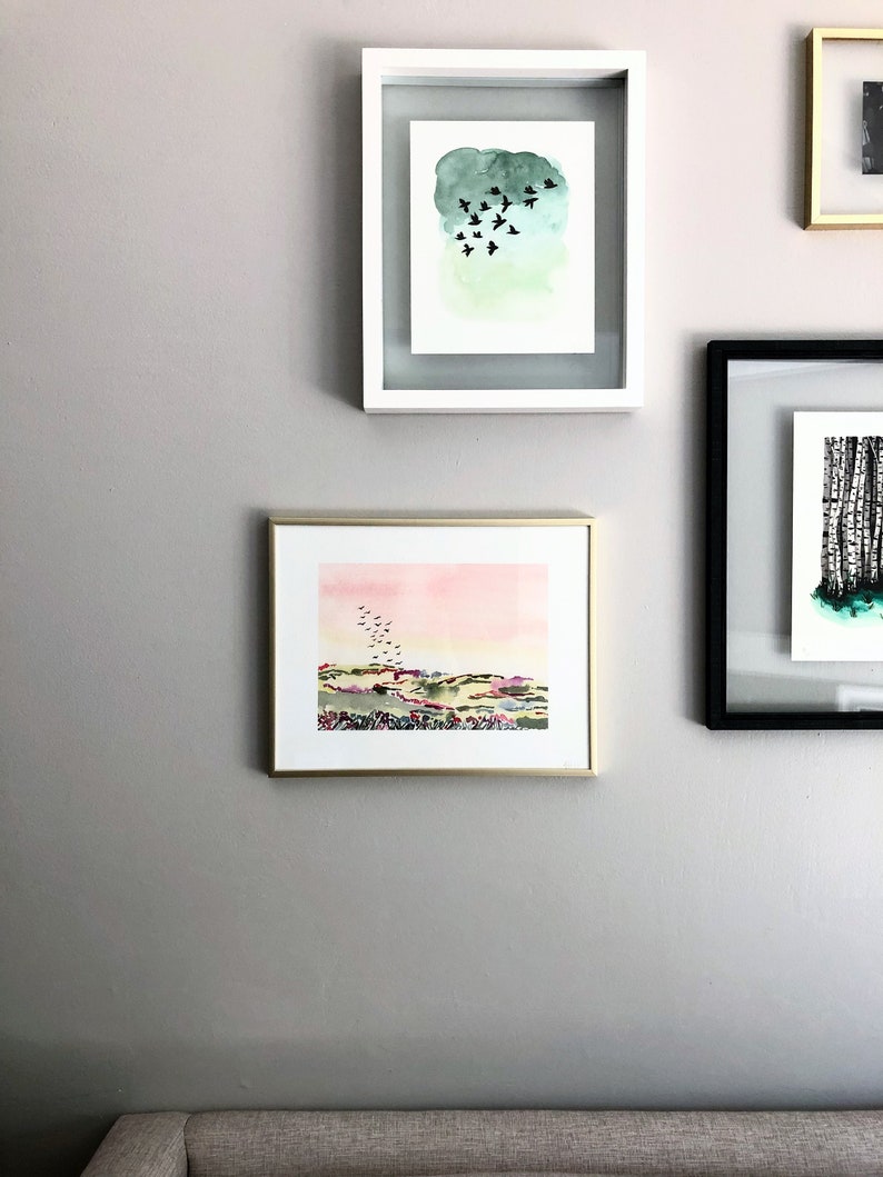 Puede incluir: Tres pinturas de acuarela enmarcadas en una pared gris claro. La pintura superior presenta un fondo de acuarela verde con una bandada de p&aacute;jaros negros volando. La pintura del medio presenta una puesta de sol de acuarela rosa y amarilla sobre un paisaje verde con una bandada de p&aacute;jaros negros volando. La pintura inferior no es visible en la imagen.