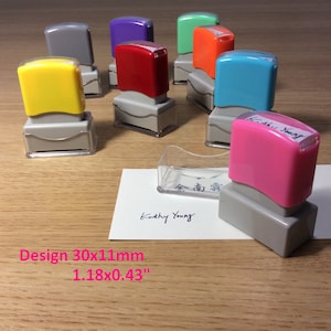 May include: Assortment of colorful self-inking stamps with gray bases. The stamps are arranged in a semi-circle on a wooden surface. The stamps are 1.18 inches by 0.43 inches. The text "Kathy Young" is written on a white card in black ink.