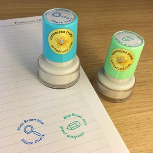 May include: Two blue and green self-inking stamps with a yellow star and smiley face. The text on the stamps reads "customized stamp artiestudio.com". The stamps are on a white lined notebook with the text "Miss Brown says" and a magnifying glass or pencil icon.