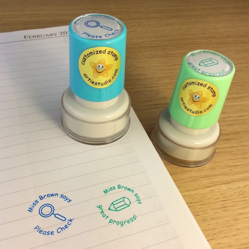 May include: Two blue and green self-inking stamps with a yellow star and smiley face. The text on the stamps reads "customized stamp artiestudio.com". The stamps are on a white lined notebook with the text "Miss Brown says" and a magnifying glass or pencil icon.