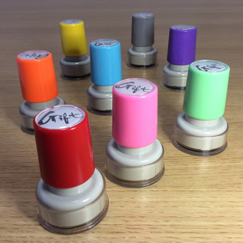 May include: Assortment of ten colorful ink pads with white lids. The ink pads are arranged in a circle on a wooden surface. The ink pads are labeled with the word "Gift" in black ink.