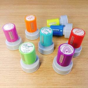 Pode incluir: Uma coleção de carimbos de borracha coloridos com vários desenhos e mensagens. Os carimbos estão sobre uma superfície de madeira. Os carimbos incluem: "You rock!" "Great Idea!" "Mrs. Hodges loves..." "Aufgabe lesen!" "Schöner arbeiten!" e "Yindemei"