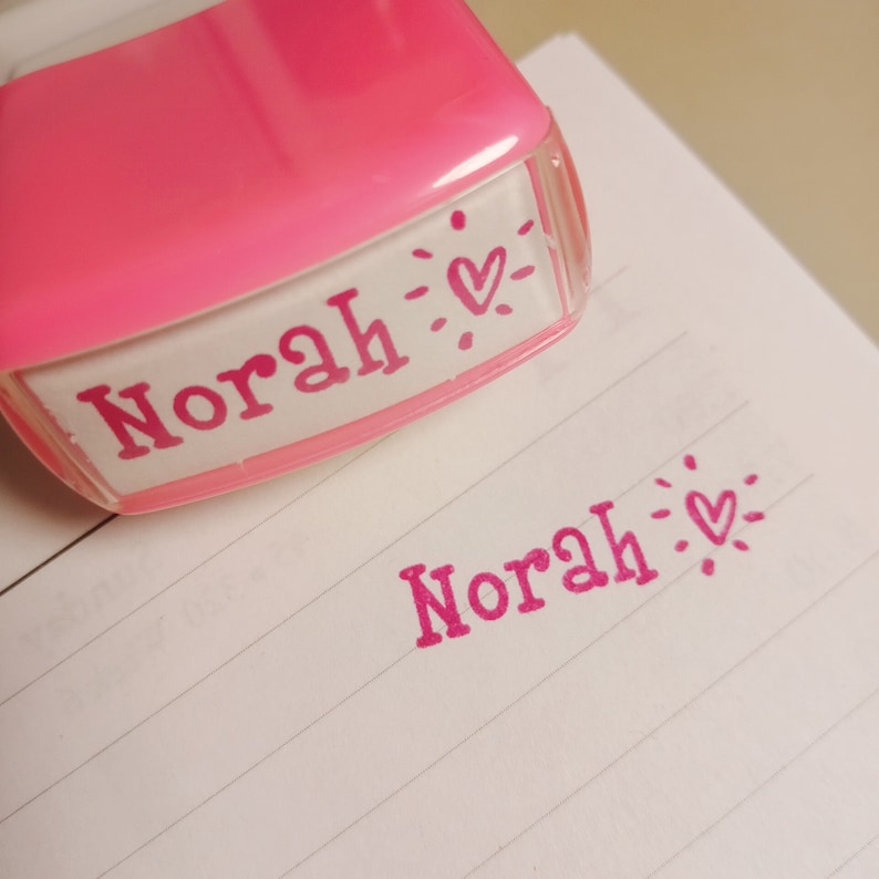 May include: Pink plastic stamp with the name "Norah" and a small heart design. The stamp is used to create a pink imprint of the name on a white lined paper.