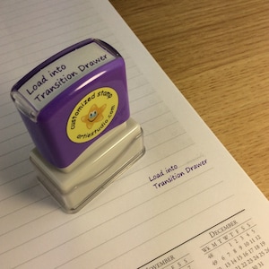 May include: Purple rubber stamp with the text "Load into Transition Drawer" and a yellow star with a smiley face. The stamp is on a lined notebook page with the text "Load into Transition Drawer" written in purple ink.