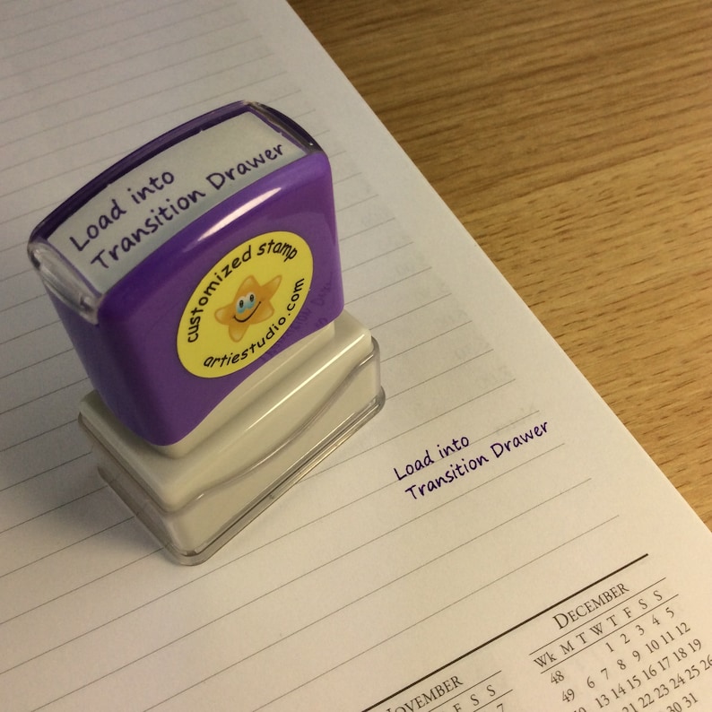 May include: Purple rubber stamp with the text "Load into Transition Drawer" and a yellow star with a smiley face. The stamp is on a lined notebook page with the text "Load into Transition Drawer" written in purple ink.