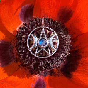 May include: Silver pendant featuring a triple moon symbol with a pentagram inside and a blue iridescent stone in the center. The pendant is resting on a red poppy flower.