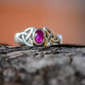 Könnte beinhalten: Ein silberner Ring mit einem rosafarbenen Edelstein, der in einem keltischen Knotendesign eingefasst ist.