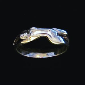 Pode incluir: Um anel de prata em forma de figura humana reclinada. O anel tem um acabamento polido e está sobre um fundo preto. A forma da figura é detalhada, com curvas e contornos visíveis.