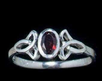 May include: A silver ring with a central oval-cut, deep red gemstone. The ring features Celtic knot designs on either side of the gemstone. The band is smooth and polished, set against a black background.