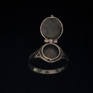 Op de afbeelding: Een zilveren ring met een scharnierend, rond medaillon dat opengaat om een lege ruimte binnenin te onthullen. De ring heeft een ingewikkeld ontwerp rond het medaillon.